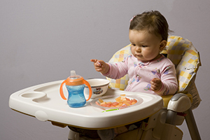 Baby sitting in high chair with sippy cup, reaching for finger foods.